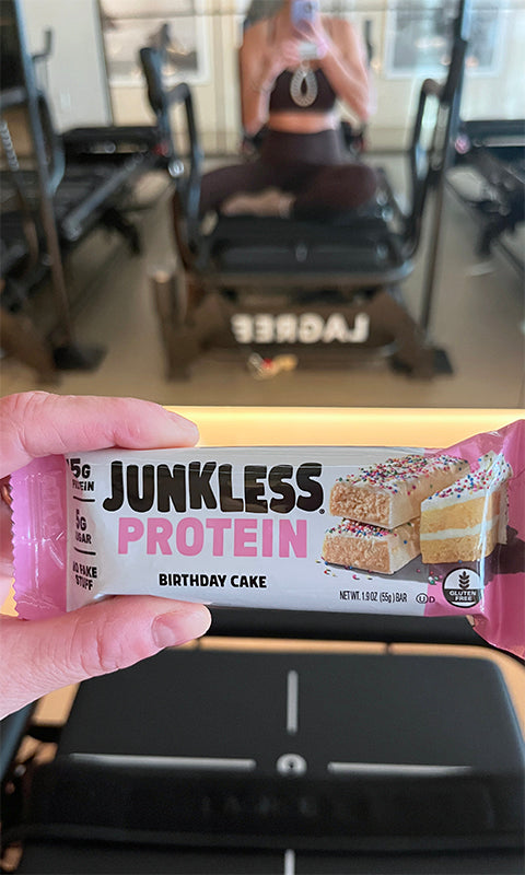 Protein bar with 'Junkless Protein' packaging held in front of a gym setting.