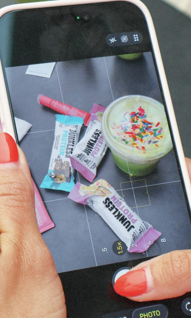 matcha next to lip gloss and junkless protein bars on a table in the screen of a phone taking a photo