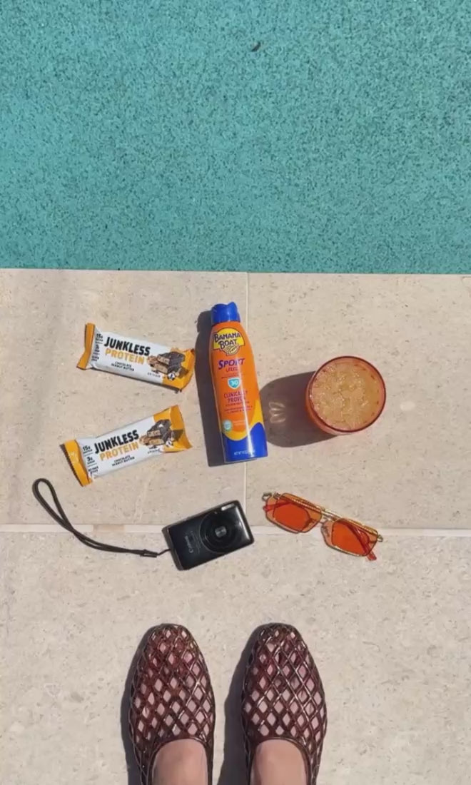 junkless protein bars next to sunscreen, a drink, camera and sunglasses in front of shoes on a sidewalk.