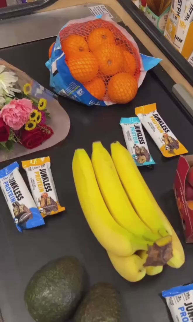 Junkless protein bars on a check out belt with bananas, oranges, flowers and avocados.